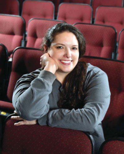 A woman smiles at the camera while sitting in a red Elbert Theatre seat with her chin resting on her hand.