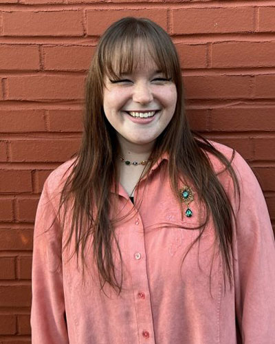 A smiling young person with long brown hair, wearing a pink button-down shirt and a green gemstone brooch, is someone to contact at the Elbert Theatre.