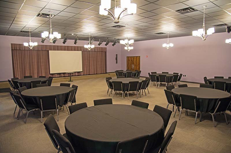 A large, empty event room with several round tables covered in black tablecloths and a projection screen in the front, set up for Elbert Theatre rentals.