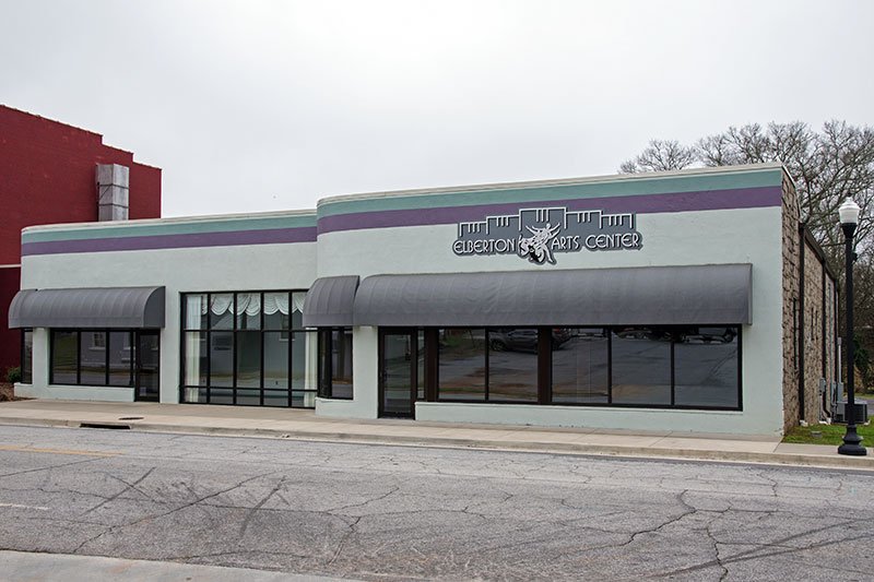 The single-story, light-colored Elberton Arts Center building features large storefront windows and dark awnings, serving as a location for Elbert Theatre rentals along the street.
