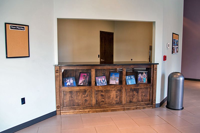 A wooden counter with a row of framed photographs sits beneath a service window at the Elbert Theatre, with a “Now Playing” corkboard on the left wall and a trash can on the right.