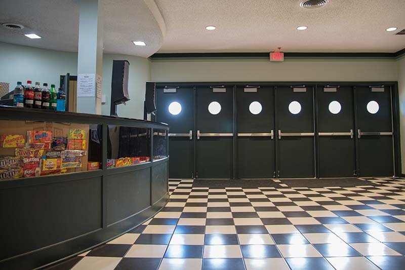 A movie theater concession stand with a glass display of candy, next to a hallway with a black and white checkered floor and a row of doors with circular windows at the Elbert Theatre rentals.