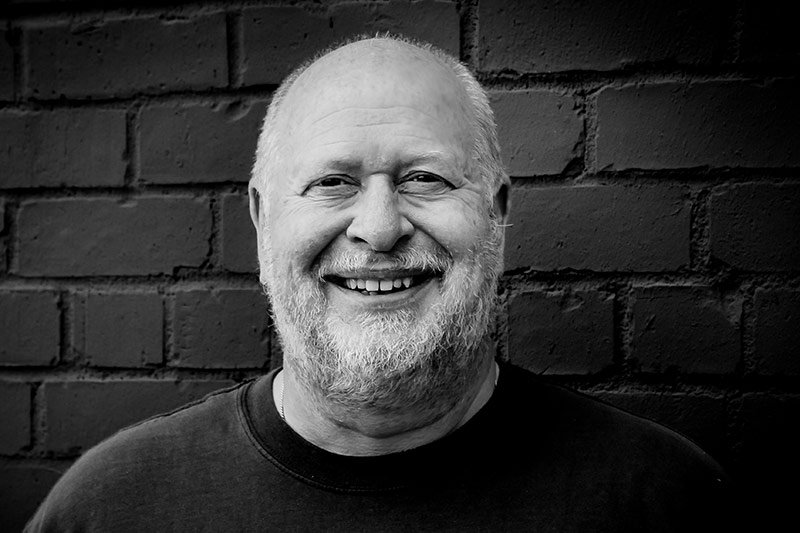 Black and white portrait of a smiling middle-aged man with a beard, positioned in front of an Elbert Theatre Foundation brick wall.