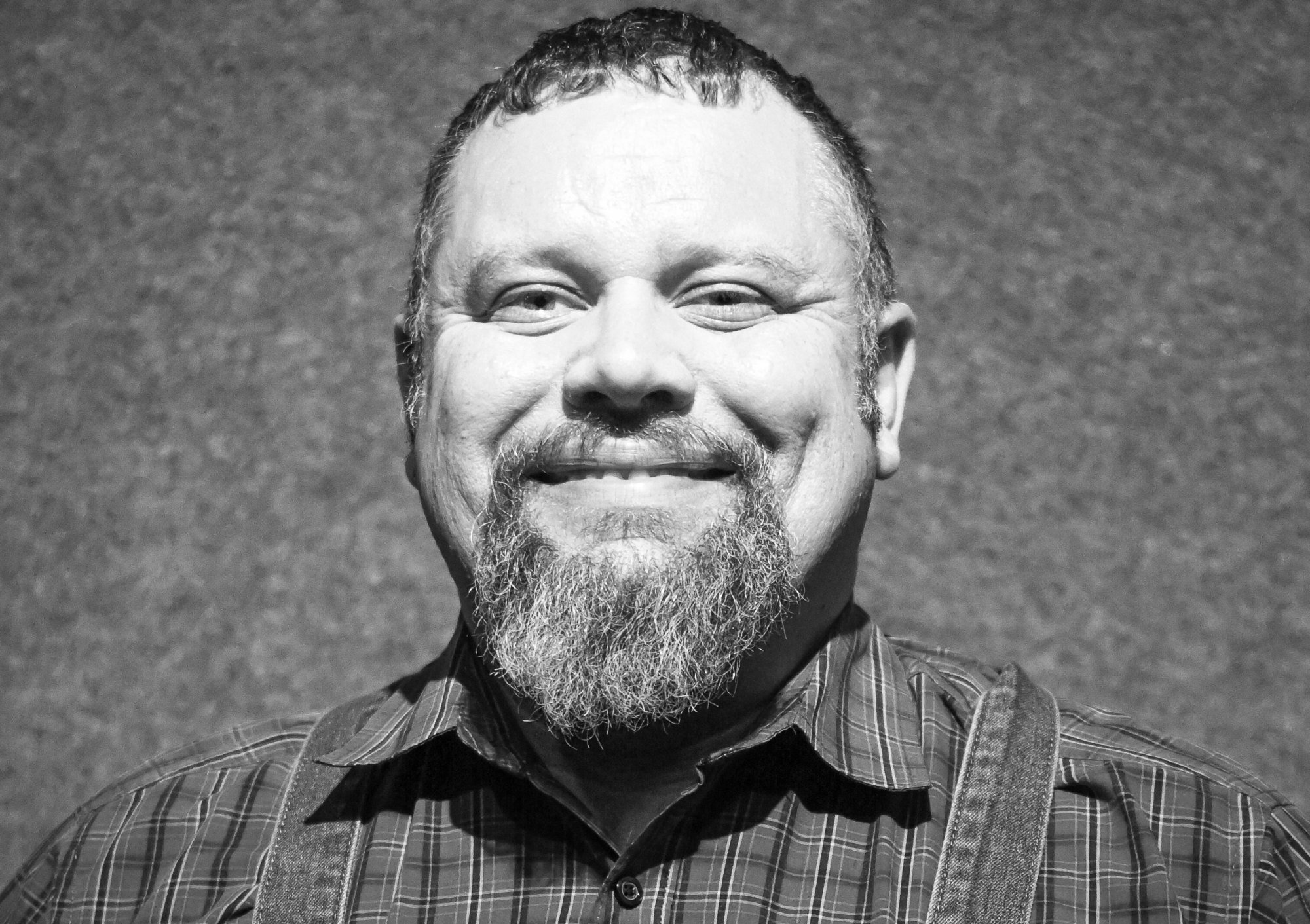 Black and white headshot of a smiling man with a goatee wearing a plaid shirt and overalls for the Elbert Theatre Foundation.