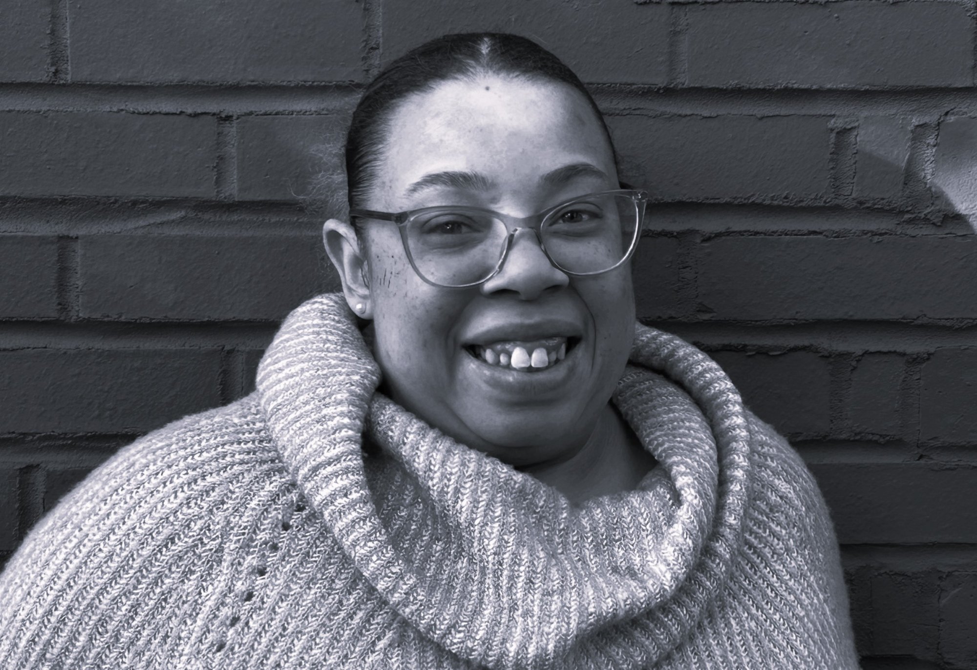 A smiling woman wearing glasses and a cowl-neck sweater stands in front of a brick wall at the Elbert Theatre Foundation.