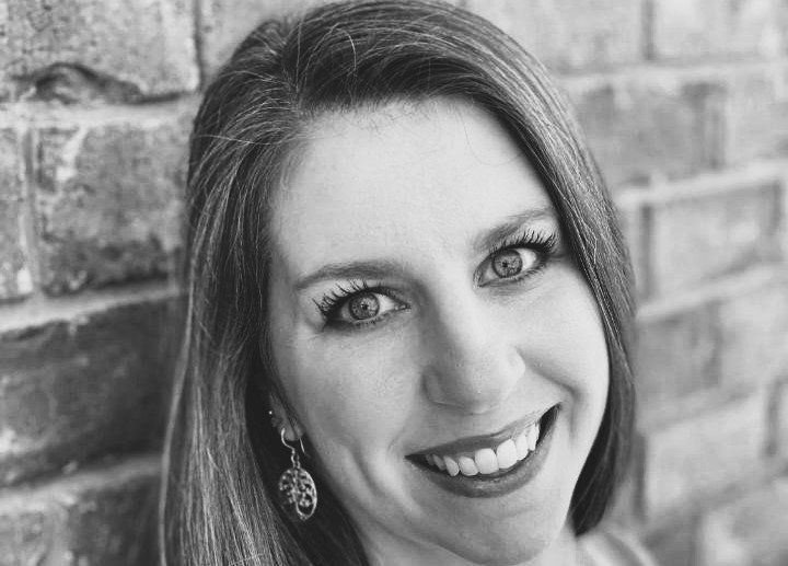 Black and white portrait of a smiling woman leaning against an Elbert Theatre Foundation brick wall.